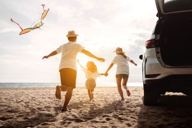 Rear view of a woman and man with their daughter running on a sunny beach away from their car while flying a kite