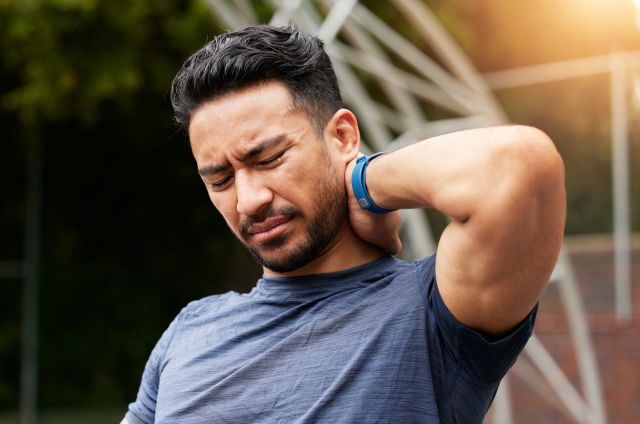 Closeup of a mid-thirties man in athletic clothing outdoors rubs his shoulder due to muscle tenderness caused by fibromyalgia