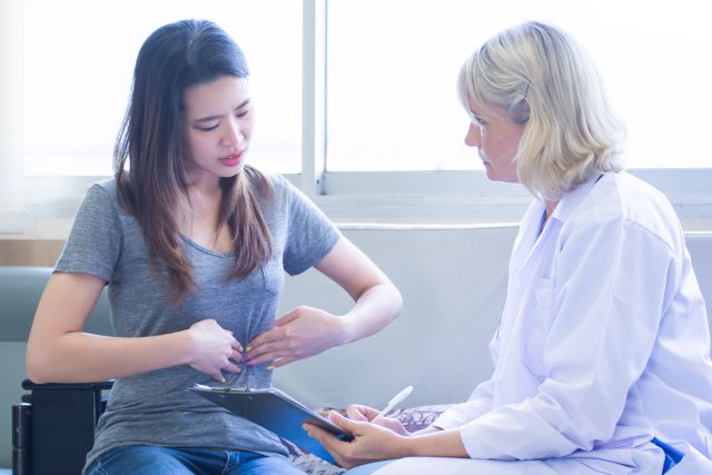 a white female doctor with white hair examines a young Asian patient who appears to be experiencing abdominal pain