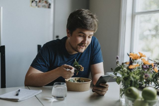 a bearded young man sits in his kitchen reading his cell phone and eating a healthy salad for lunch