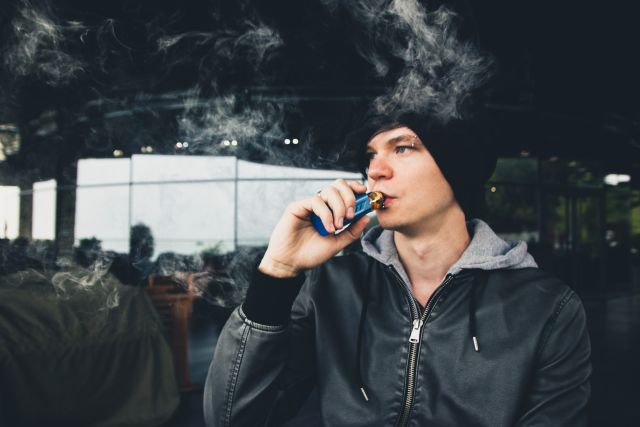 a white teenage boy wearing a knit hat and an eyebrow piercing uses a vaping or e-cigarette device 