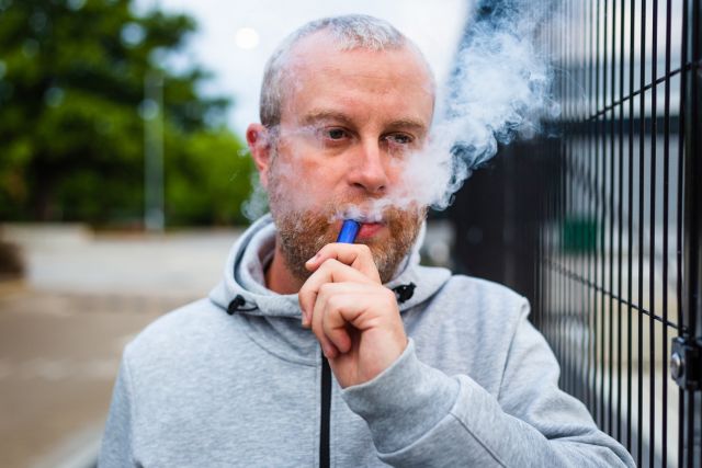 a middle aged white man with a beard stands outdoors puffing smoke from a vape or e-cigarette