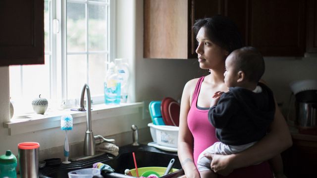 Woman feeling overwhelmed holds a young baby in a messy kitchen 
