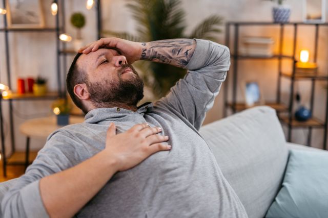 A slightly overweight white man with a tattoo on his arm sits on his coach, leaning back, holding his hand to his head. He feels dizzy and has a headache. Could he be having a stroke?