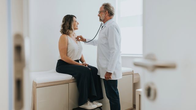 A healthcare provider uses a stethoscope to listen to the heart of a middle-aged woman during an appointment.