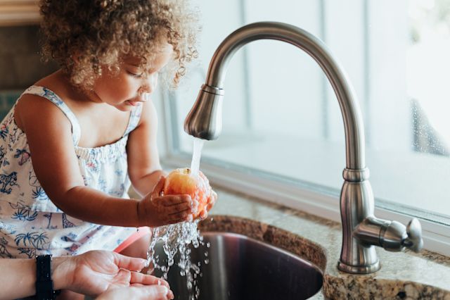 a young Black girl washes a piece of fruit under running water in her family's kitchen sink, with help from a parent