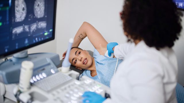 A healthcare provider checks a female patient's lymph nodes with an ultrasound. Lymph nodes in the chest, armpit, and around the collarbone are a potential site for a breast cancer recurrence.