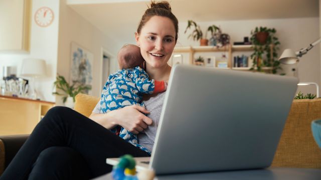 A new mom holds her baby in one arm as she uses a computer to research achondroplasia organizations.