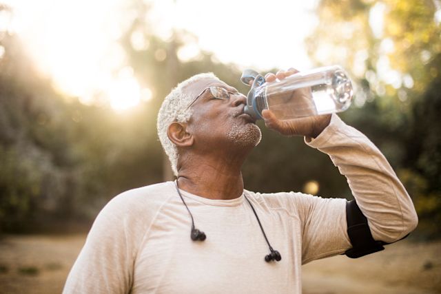 An older Black man wearing exercise gear takes a sip from a bottle of water to hydrate after working out outdoors