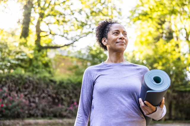 Black adult female in workout clothing holding a rolled up yoga mat outside