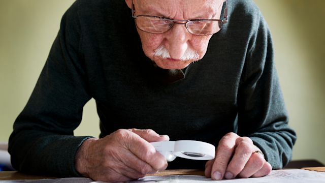 An elderly man uses a magnifying glass to aid him in reading a book.