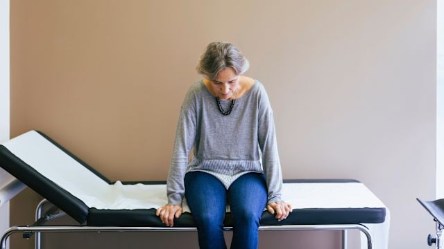 A woman sits on a bed in an exam room waiting to talk to her healthcare provider about cancer fatigue symptoms.