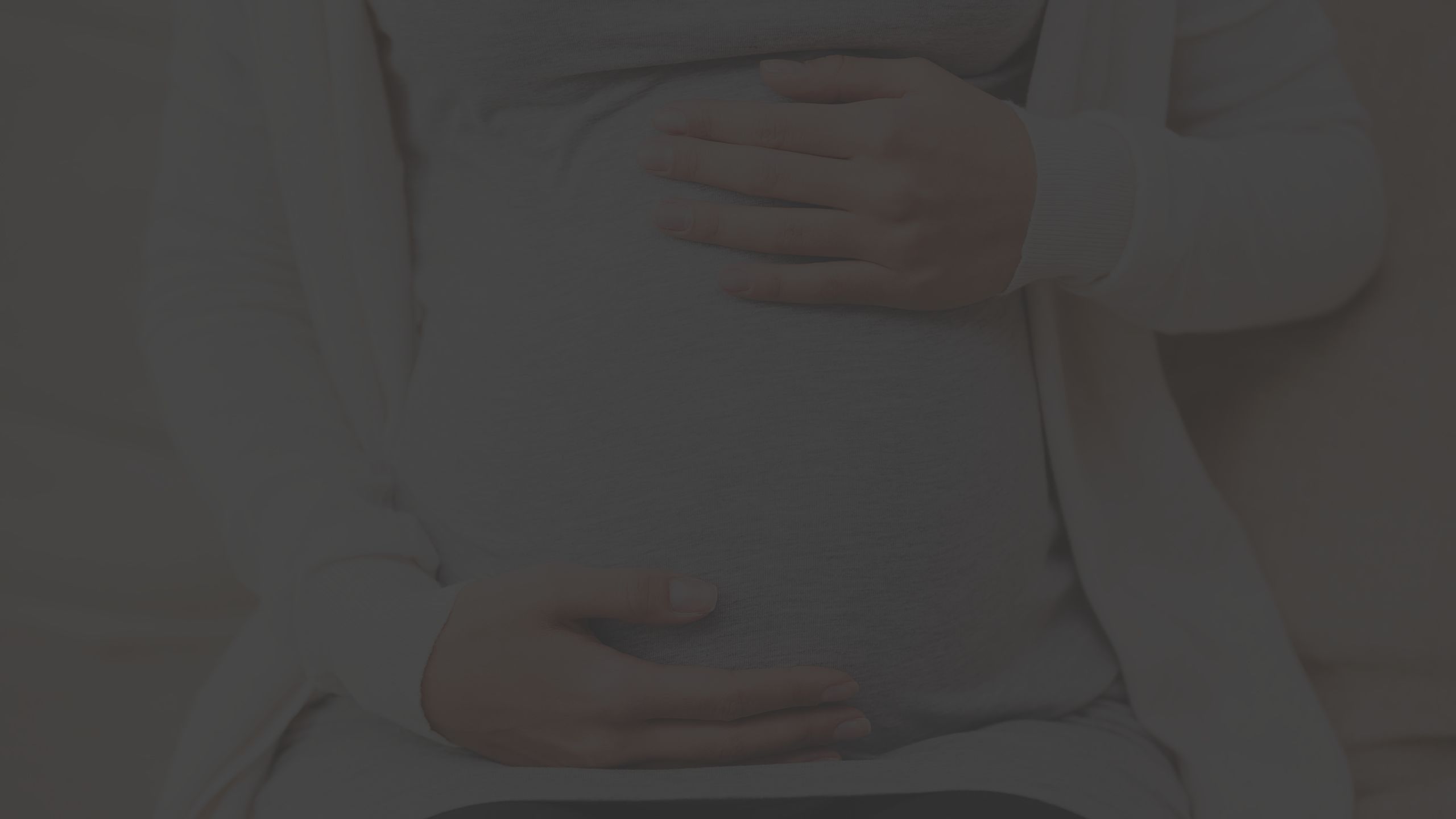 pregnant person dressed in a light gray shirt and cream colored sweater, sitting down and holding their belly