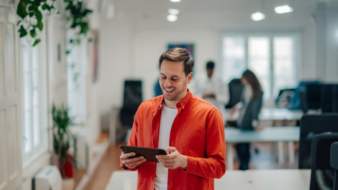 A smiling man in a red shirt holds a tablet in an office.
