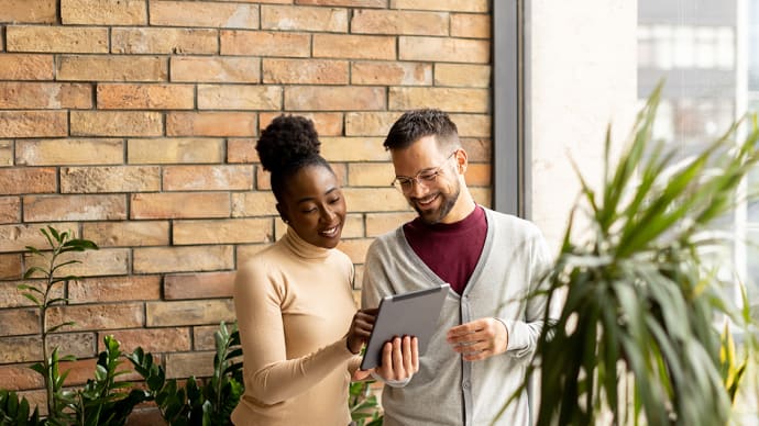 Two smiling colleagues, a Black woman and a white man, look at a tablet together in an office with a brick wall.